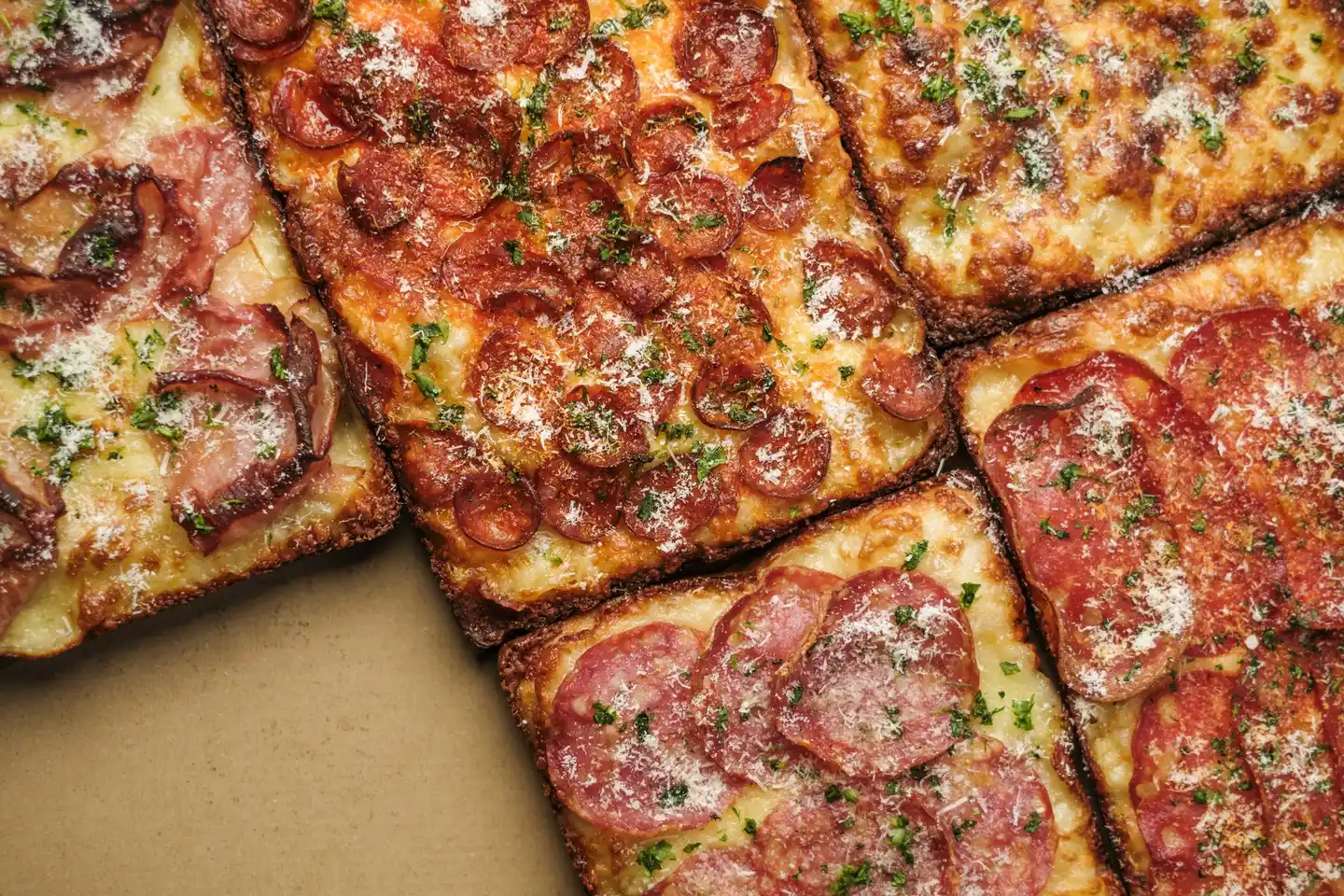 Szalámis, kolbászos és sonkás pizzák válogatása, sajttal és friss zöld fűszerrel megszórva, kartondobozban tálalva.